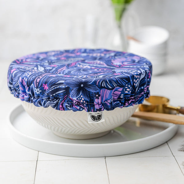 White bowl with a blue floral lid on a white surface, with a blurred background of flowers and kitchen items.