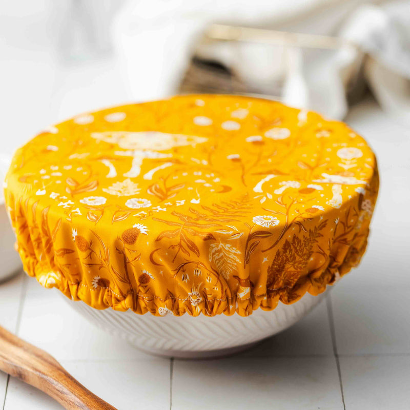 Yellow floral-patterned fabric cover on a white bowl with a wooden tool on a light surface.