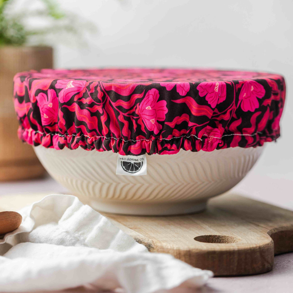 White bowl with a pink floral cover on a wooden cutting board, with a plant and cloth in the background.
